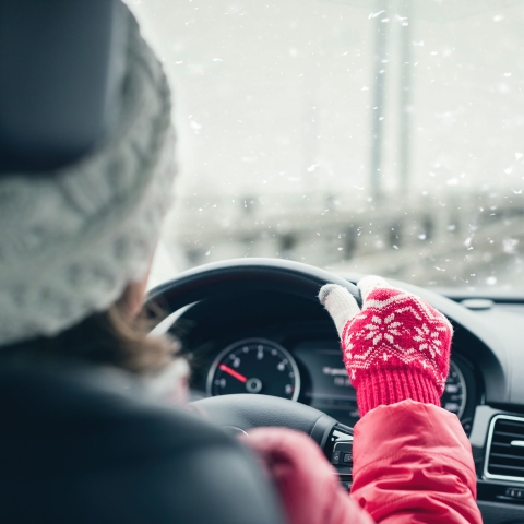 Woman in red jacket, hat, and gloves driving on highway during winter snowfall