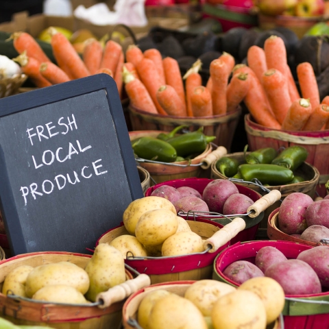 Fresh Local Produce sign at a farmer's market surrounded by potatoes, carrots, and other vegetables