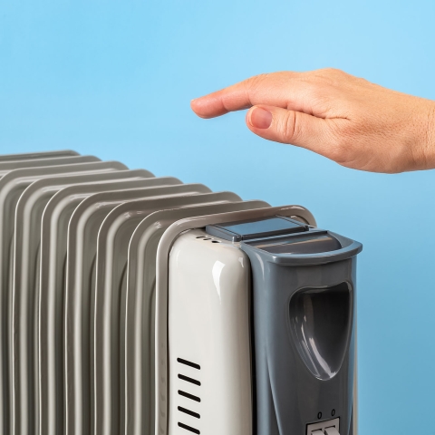 The top of a silver space heater with a hand over it feeling the heat against a light blue wall