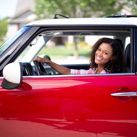 A woman in a red car with her hand on the steering wheel