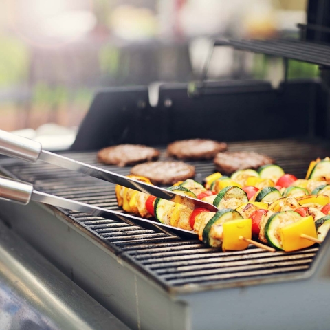 A hand holding a set of tongs turning a vegetable skewer on a bbq grill with burgers in the background