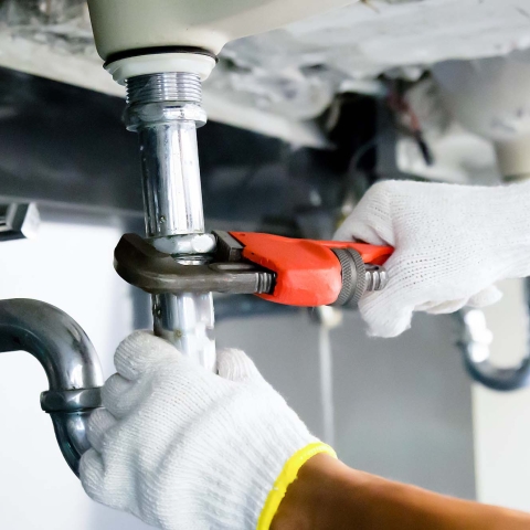 Two hands wearing white gloves working on silver plumbing fixtures underneath a bathroom sink