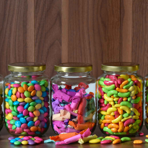 Five clear glass jars with gold lids arranged in a row on a wooden surface, each filled with different colorful candies, including gummy drops, candy-coated chocolates, wrapped sweets, jelly beans, and fruit-shaped candies, with a few pieces scattered in front.