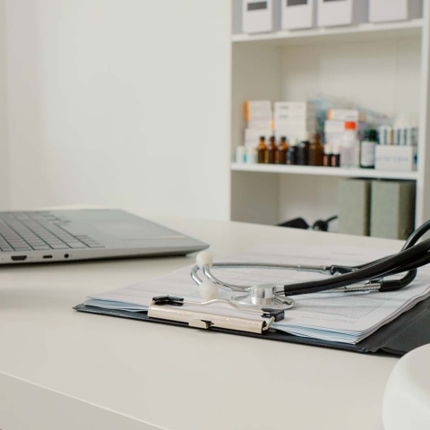 A clean medical office desk with a stethoscope resting on a clipboard of documents, a laptop and small potted plant nearby, and shelves with medicine bottles in the background.