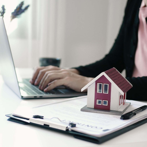Person typing on a laptop at a desk with a small model house placed on top of a clipboard with documents, suggesting real estate work