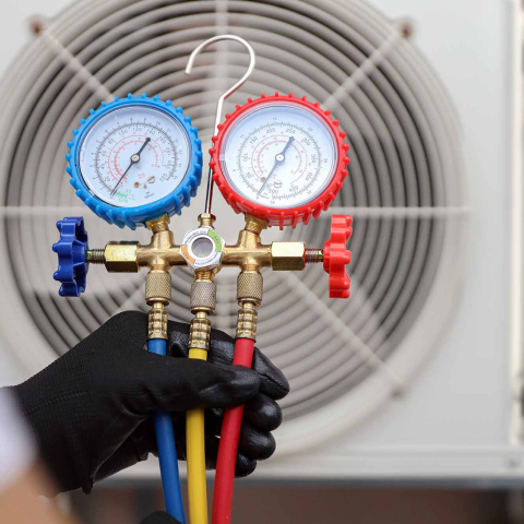 HVAC technician holding a manifold gauge set with blue, red, and yellow hoses while checking refrigerant pressure on an outdoor air conditioning condenser unit.