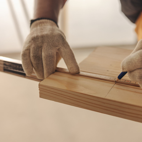 Close-up of a person wearing work gloves measuring and marking a wooden board with a ruler and pencil during a woodworking or carpentry project.