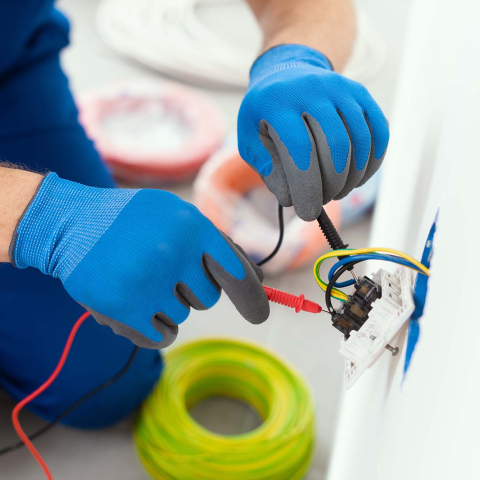Close-up of a person wearing protective gloves testing electrical wiring in a wall outlet with a multimeter, with electrical cable and installation materials visible nearby.