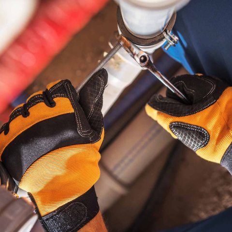 Close-up of a worker wearing orange and black protective gloves using a wrench to tighten a clamp on a white PVC pipe connection in a plumbing system