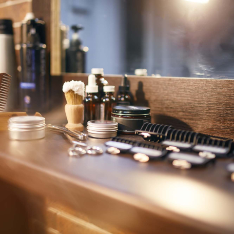 Barbering and grooming tools arranged on a wooden counter in front of a mirror, including combs, scissors, a shaving brush, clippers, and grooming products.