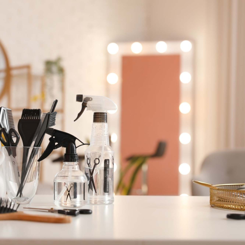 Hair styling tools and spray bottles arranged on a salon workstation, with brushes, combs, scissors, and illuminated mirrors in the background.
