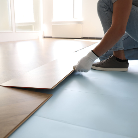 A person wearing work gloves installs wood-look flooring planks in a bright, sunlit room.
