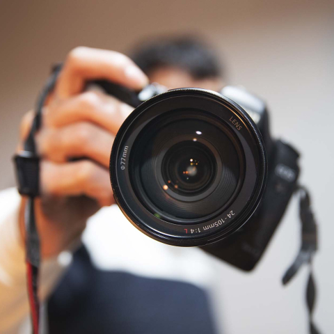Close-up of a photographer holding a camera toward the viewer, with the large camera lens in sharp focus.