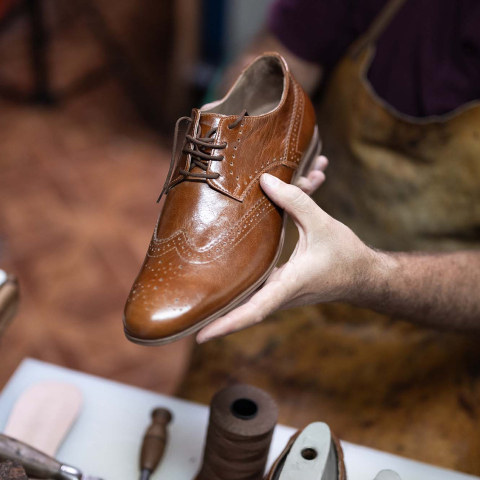 Shoe repair professional holding a polished brown leather dress shoe above a workbench with repair tools and materials.