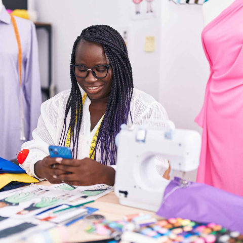 Smiling tailor reviews her phone at a worktable with a sewing machine, fabric swatches, thread, and garments.