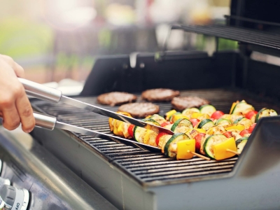 A hand holding a set of tongs turning a vegetable skewer on a bbq grill with burgers in the background