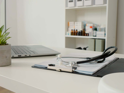 A clean medical office desk with a stethoscope resting on a clipboard of documents, a laptop and small potted plant nearby, and shelves with medicine bottles in the background.
