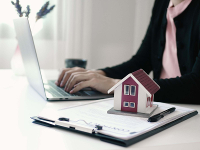 Person typing on a laptop at a desk with a small model house placed on top of a clipboard with documents, suggesting real estate work