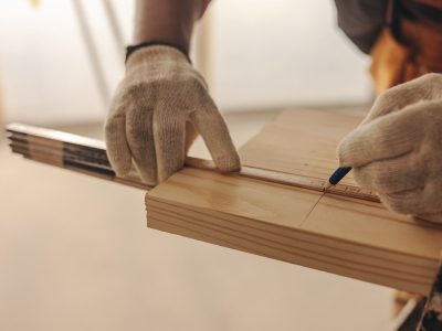 Close-up of a person wearing work gloves measuring and marking a wooden board with a ruler and pencil during a woodworking or carpentry project.
