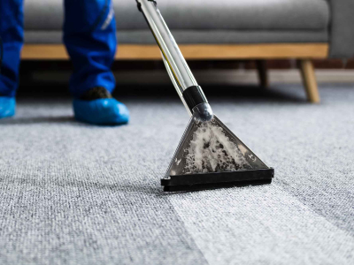 Professional carpet cleaning in progress using a hot water extraction wand on a gray carpet, with a technician wearing blue protective shoe covers visible in the background.