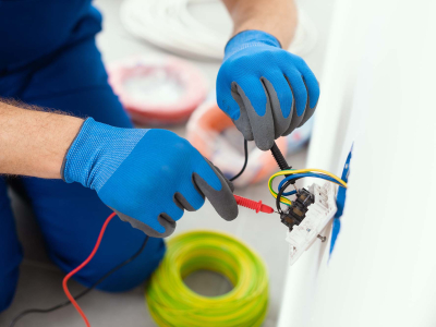 Close-up of a person wearing protective gloves testing electrical wiring in a wall outlet with a multimeter, with electrical cable and installation materials visible nearby.