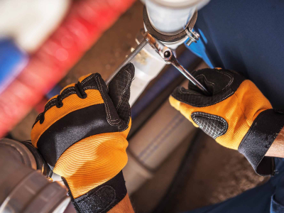 Close-up of a worker wearing orange and black protective gloves using a wrench to tighten a clamp on a white PVC pipe connection in a plumbing system