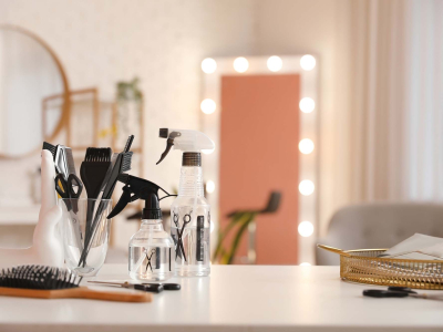 Hair styling tools and spray bottles arranged on a salon workstation, with brushes, combs, scissors, and illuminated mirrors in the background.