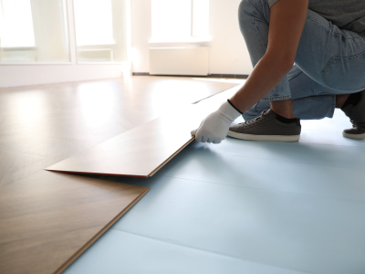 A person wearing work gloves installs wood-look flooring planks in a bright, sunlit room.