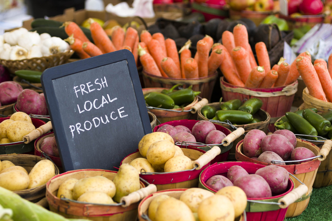 Fresh Local Produce sign at a farmer's market surrounded by potatoes, carrots, and other vegetables