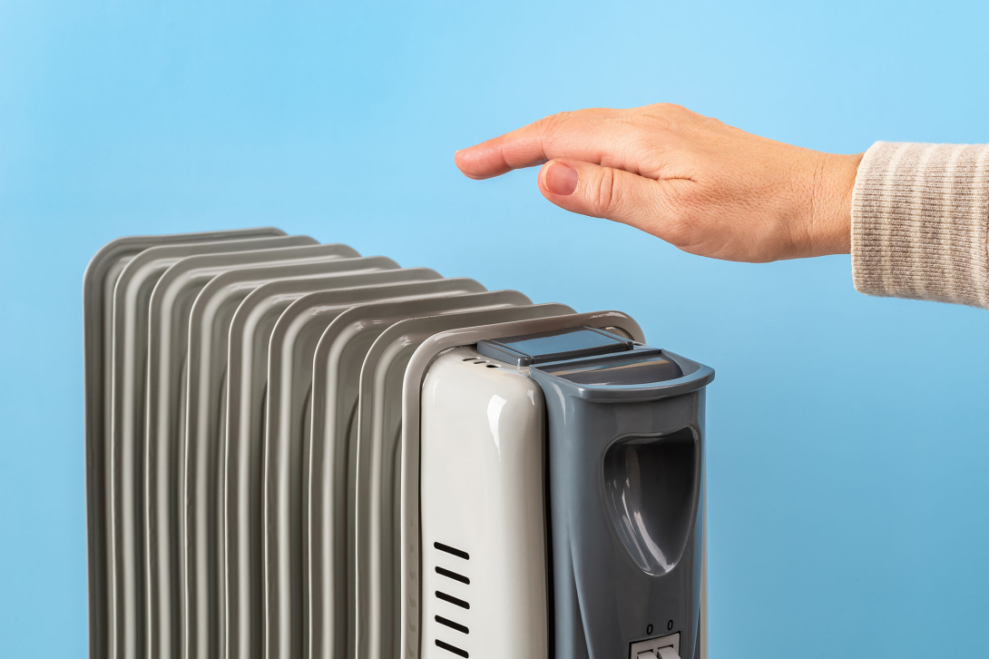 The top of a silver space heater with a hand over it feeling the heat against a light blue wall