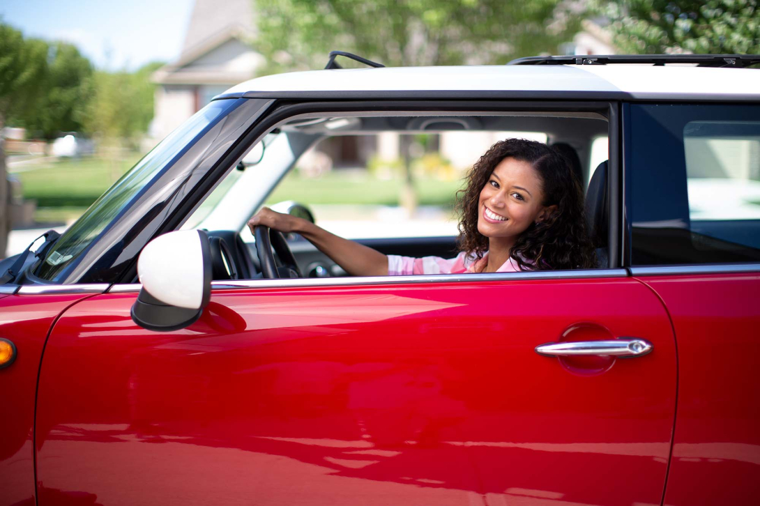 A woman in a red car with her hand on the steering wheel