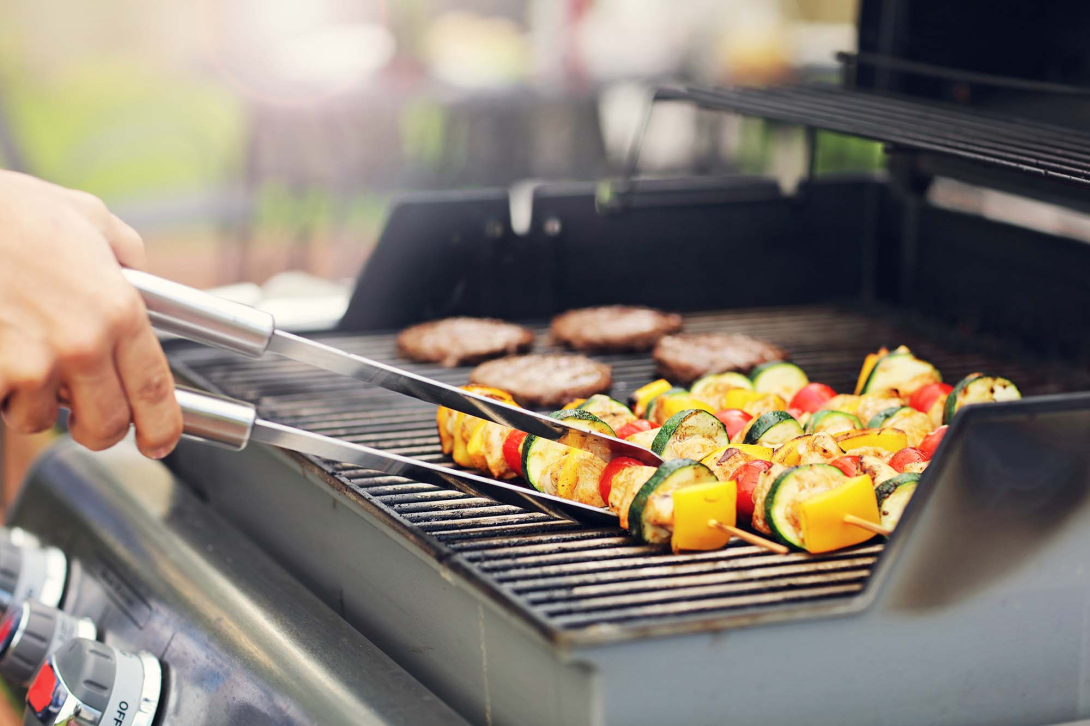 A hand holding a set of tongs turning a vegetable skewer on a bbq grill with burgers in the background