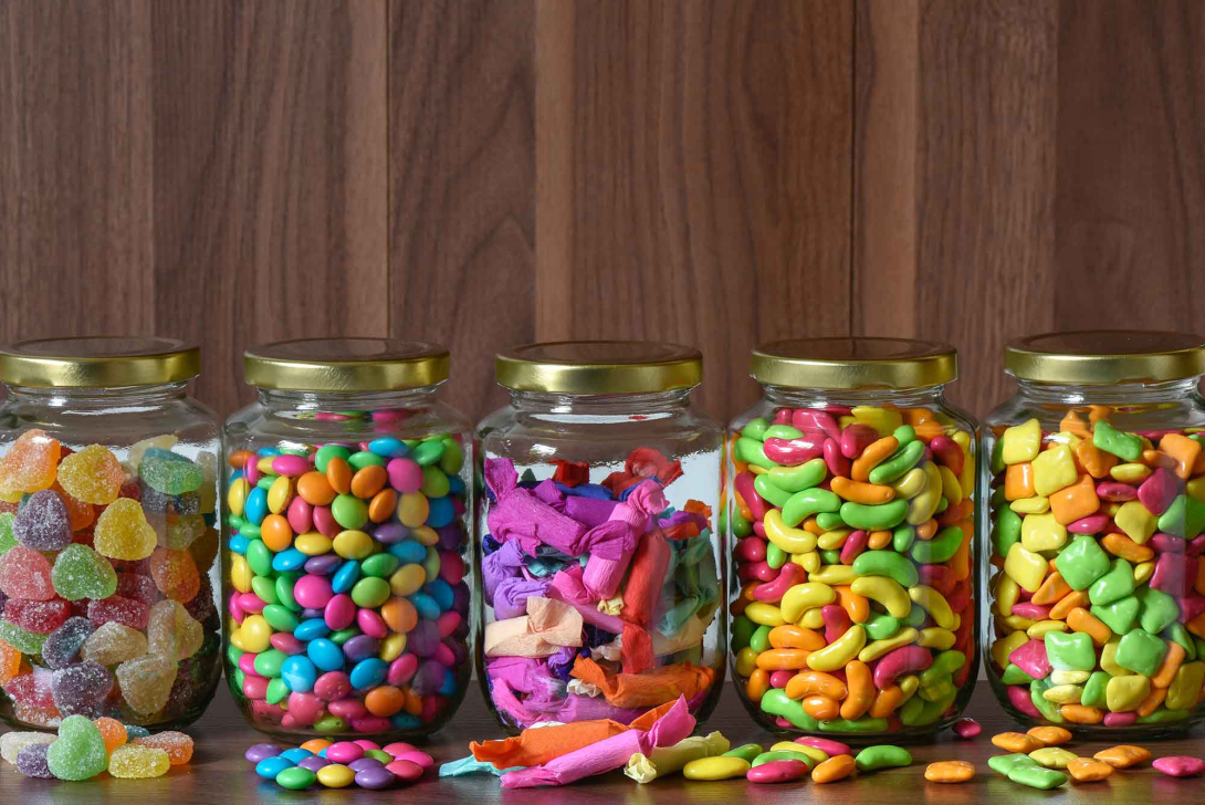 Five clear glass jars with gold lids arranged in a row on a wooden surface, each filled with different colorful candies, including gummy drops, candy-coated chocolates, wrapped sweets, jelly beans, and fruit-shaped candies, with a few pieces scattered in front.