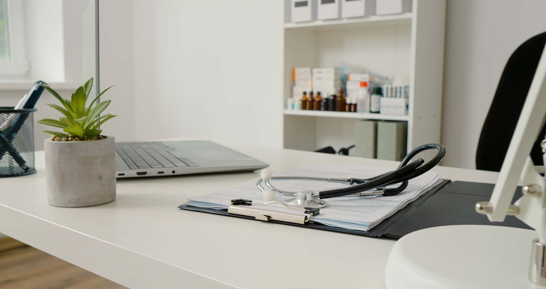A clean medical office desk with a stethoscope resting on a clipboard of documents, a laptop and small potted plant nearby, and shelves with medicine bottles in the background.