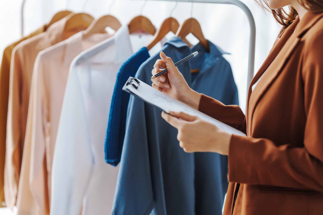 Person holding a clipboard and pen while reviewing clothing on a rack, with several garments hanging on wooden hangers in the background.