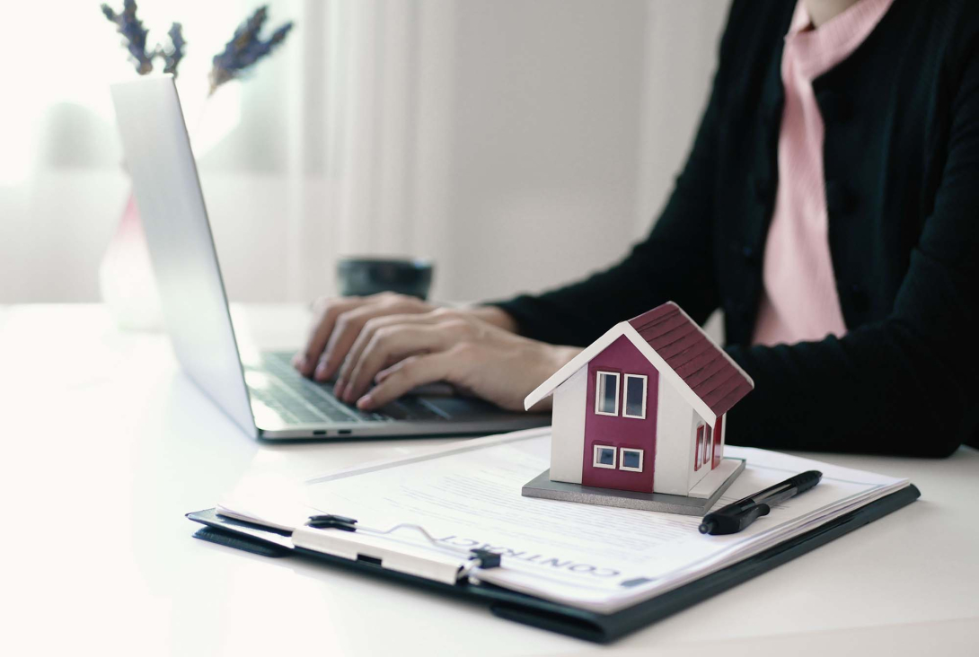 Person typing on a laptop at a desk with a small model house placed on top of a clipboard with documents, suggesting real estate work