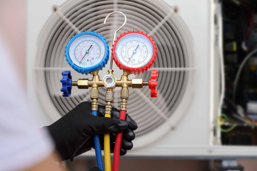 HVAC technician holding a manifold gauge set with blue, red, and yellow hoses while checking refrigerant pressure on an outdoor air conditioning condenser unit.