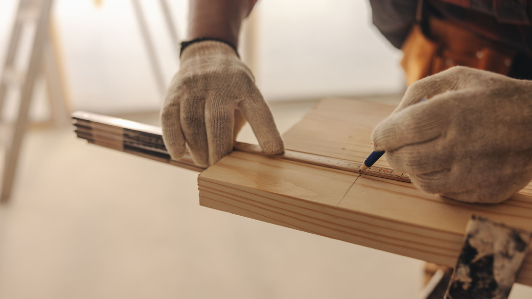 Close-up of a person wearing work gloves measuring and marking a wooden board with a ruler and pencil during a woodworking or carpentry project.