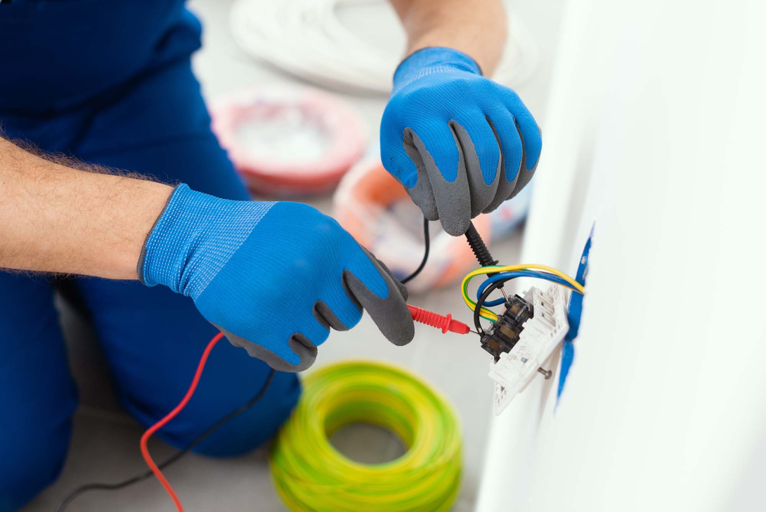 Close-up of a person wearing protective gloves testing electrical wiring in a wall outlet with a multimeter, with electrical cable and installation materials visible nearby.