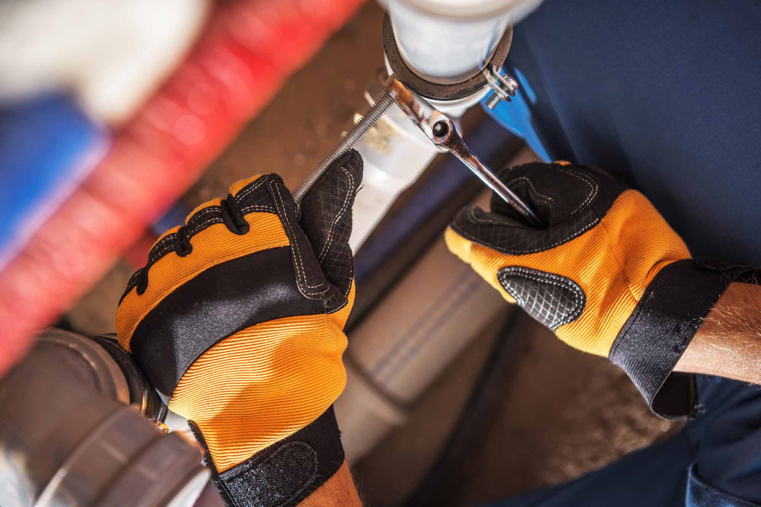 Close-up of a worker wearing orange and black protective gloves using a wrench to tighten a clamp on a white PVC pipe connection in a plumbing system