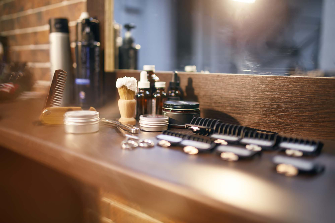 Barbering and grooming tools arranged on a wooden counter in front of a mirror, including combs, scissors, a shaving brush, clippers, and grooming products.