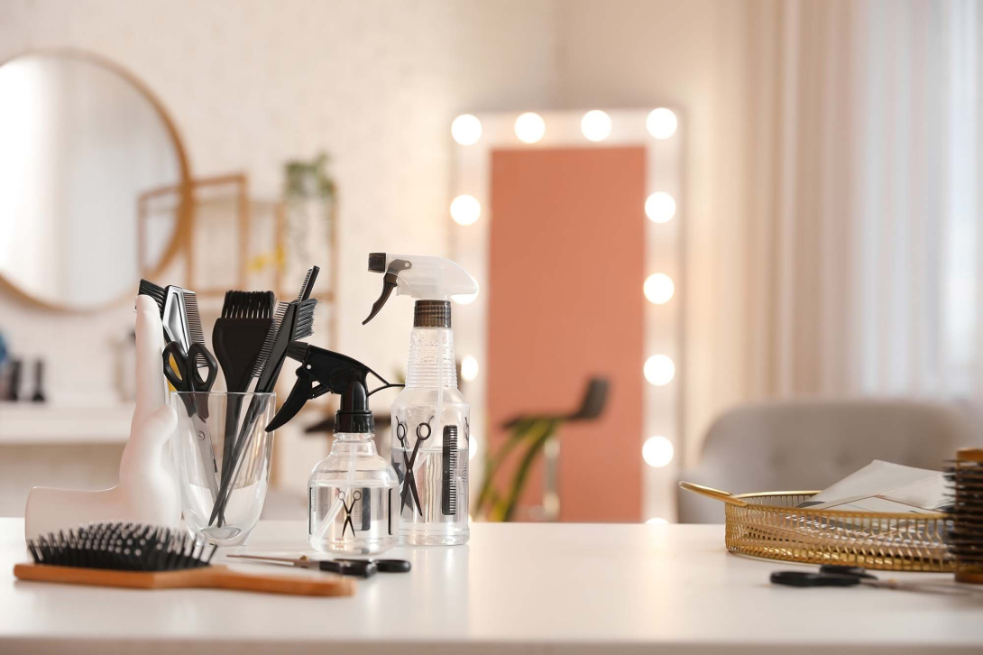 Hair styling tools and spray bottles arranged on a salon workstation, with brushes, combs, scissors, and illuminated mirrors in the background.