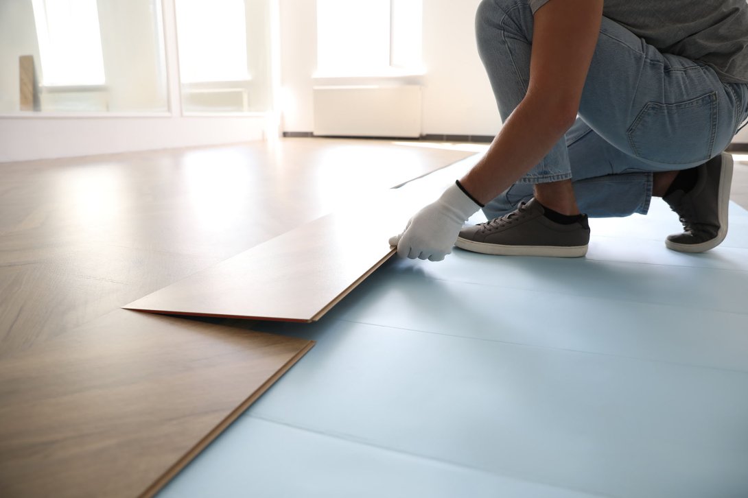 A person wearing work gloves installs wood-look flooring planks in a bright, sunlit room.
