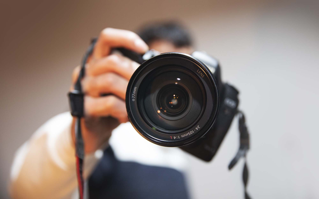 Close-up of a photographer holding a camera toward the viewer, with the large camera lens in sharp focus.