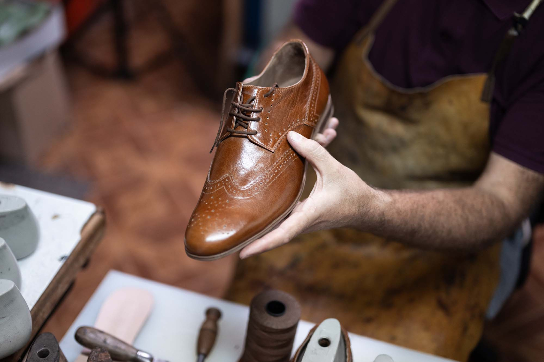Shoe repair professional holding a polished brown leather dress shoe above a workbench with repair tools and materials.
