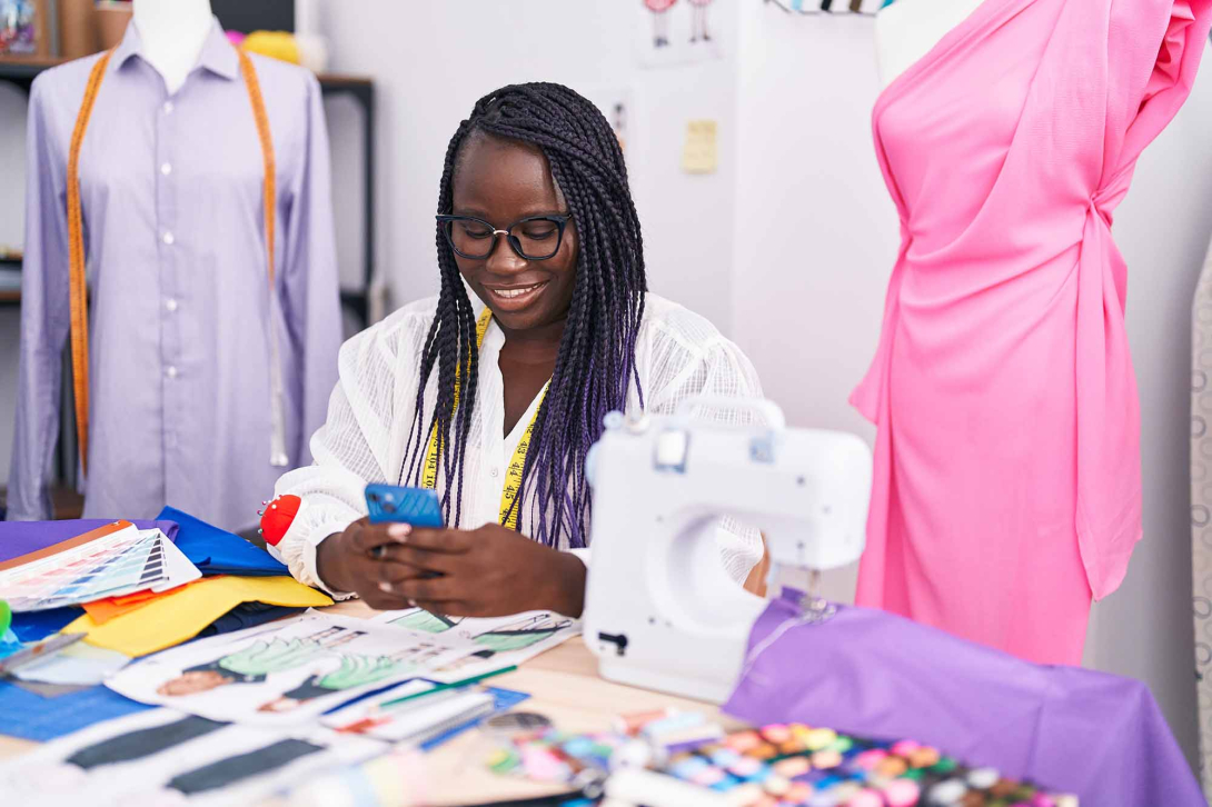 Smiling tailor reviews her phone at a worktable with a sewing machine, fabric swatches, thread, and garments.